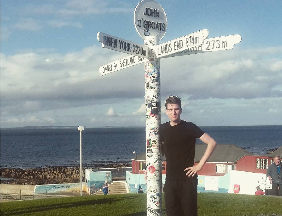 A man in a black T-Shirt standing next to the John O'Groats sign pointing to Land's End